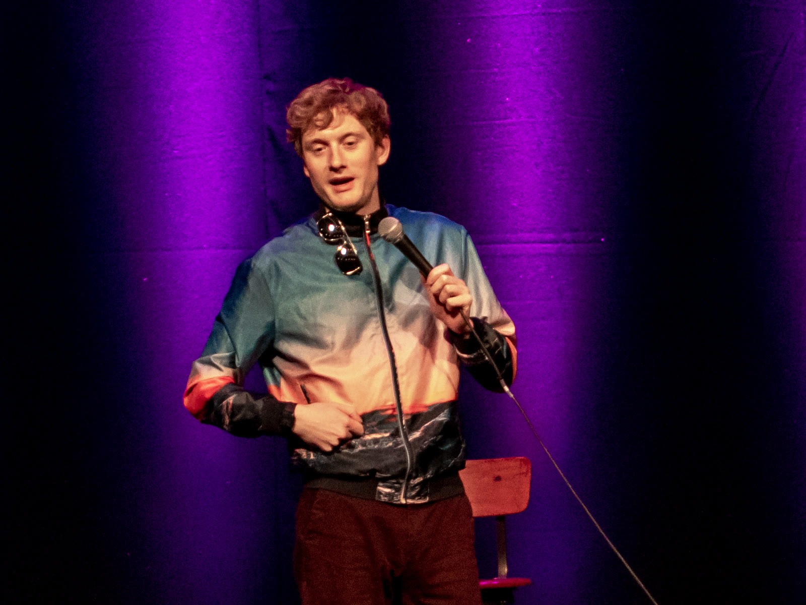 James Acaster performing stand-up comedy on stage with a microphone, wearing a colorful jacket under purple stage lighting