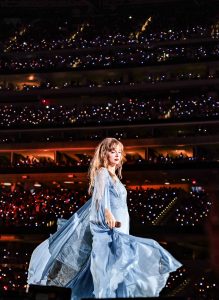 Taylor Swift performing on stage during her Eras Tour in a packed stadium at night, with thousands of fans holding up glowing phone lights