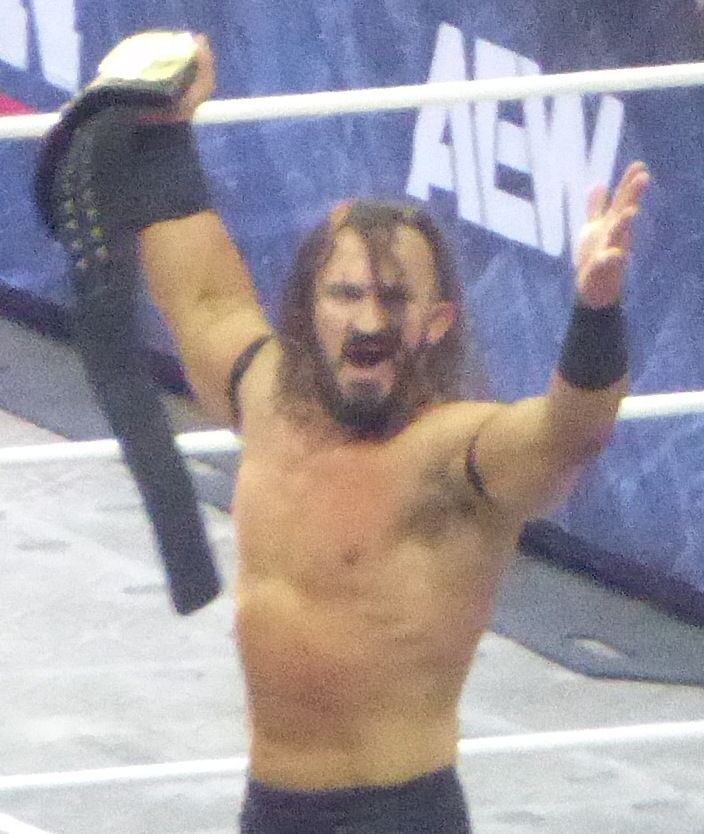 Pac celebrating with a championship belt raised in an AEW ring