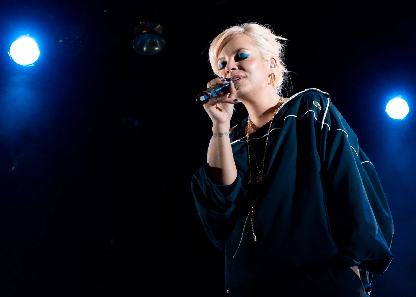 Lily Allen singing into a microphone on stage at a 2018 concert, lit by blue stage lights