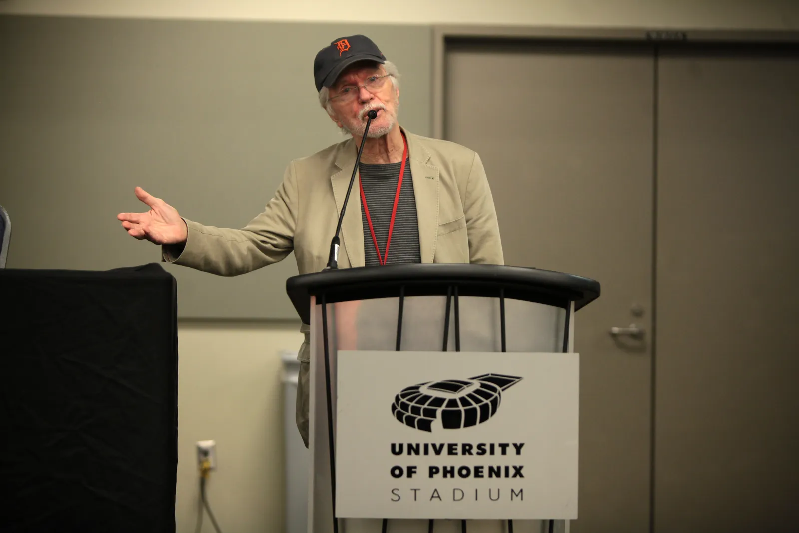 Tom Skerritt speaking at a podium at the 2014 Phoenix Comicon Fan Fest, wearing a baseball cap and glasses
