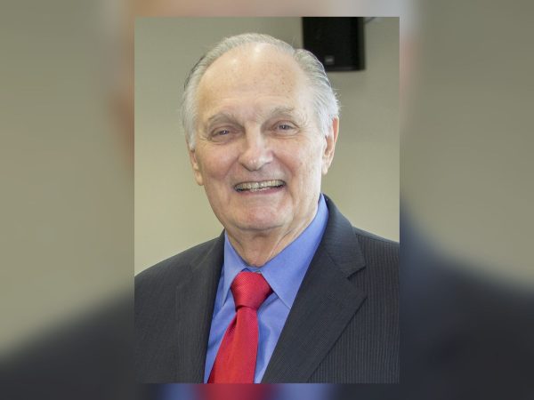 Alan Alda smiling in a dark suit and red tie, portrait from 2015