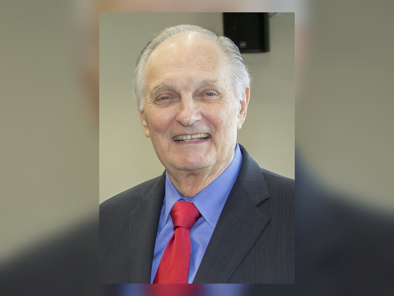 Alan Alda smiling in a dark suit and red tie, portrait from 2015
