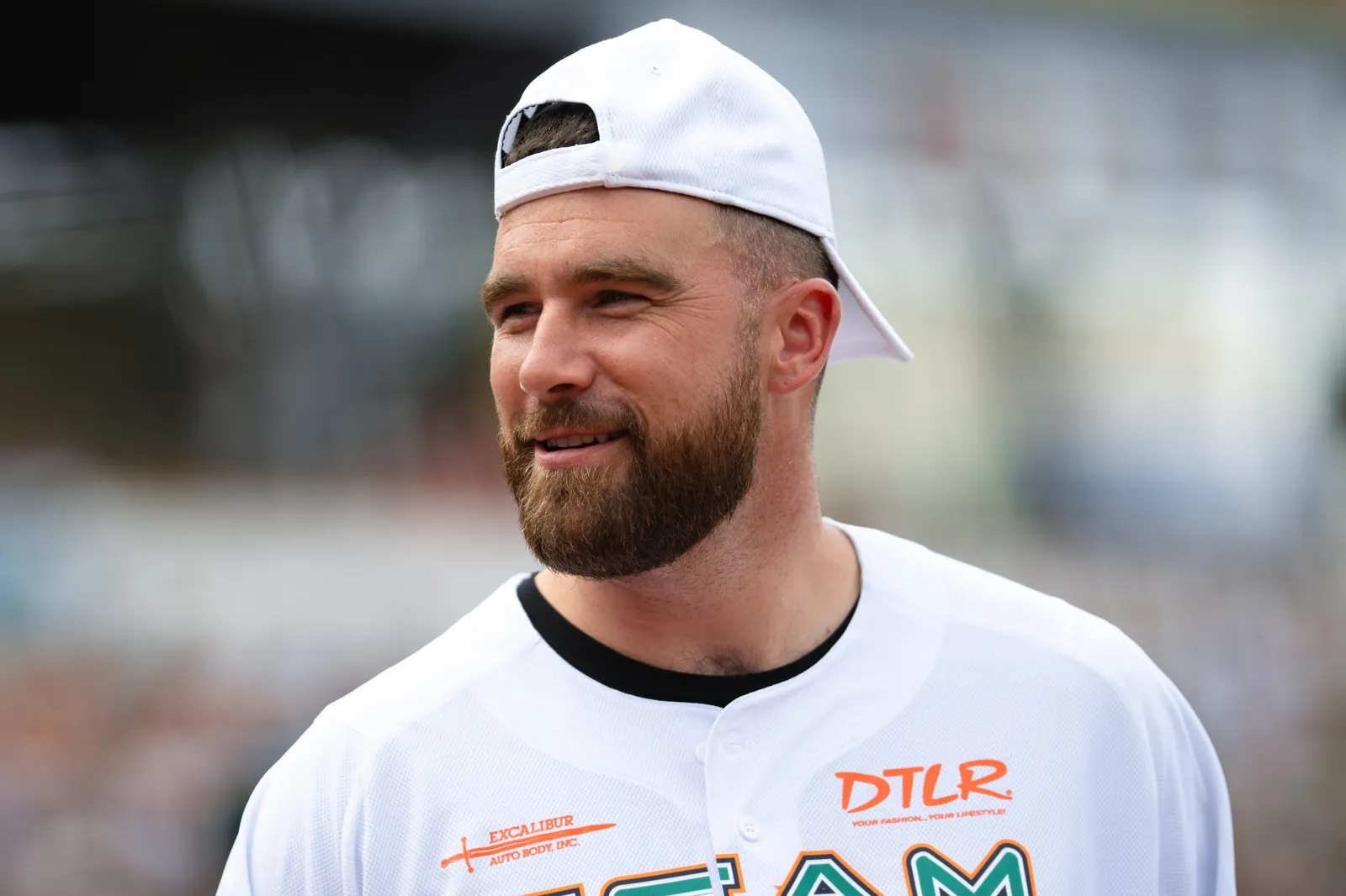 Travis Kelce wearing a white cap and jersey at a public appearance