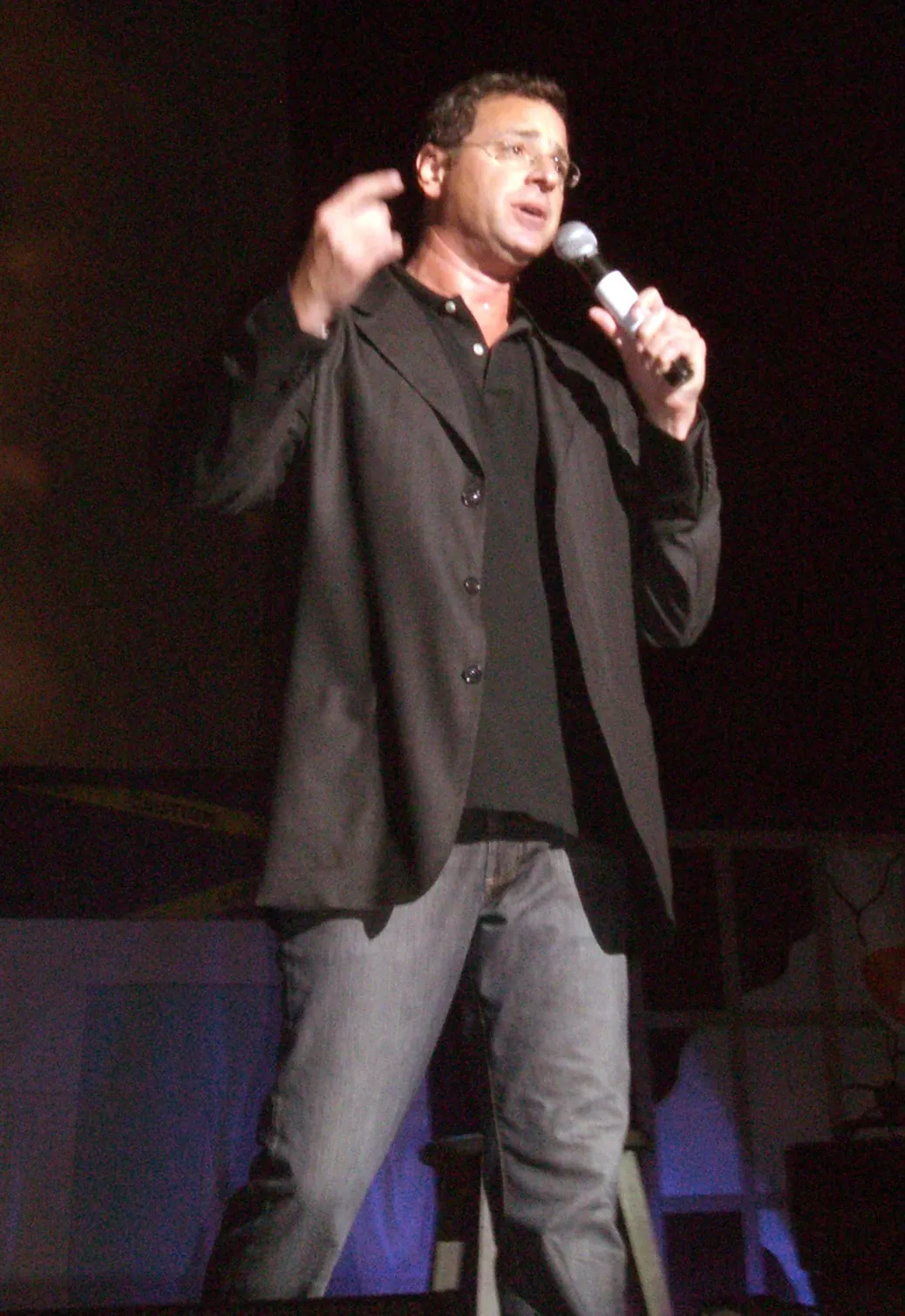 Bob Saget performing stand-up comedy on stage with a microphone