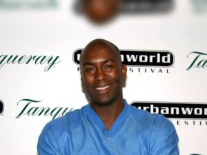 Reggie McFadden smiling in a blue shirt with arms crossed at a festival step-and-repeat backdrop.