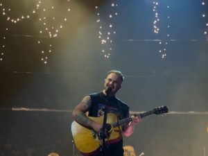 Zach Bryan performing on stage with an acoustic guitar, his full head in frame