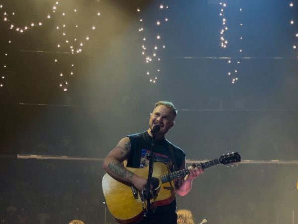 Zach Bryan performing on stage with an acoustic guitar, his full head in frame
